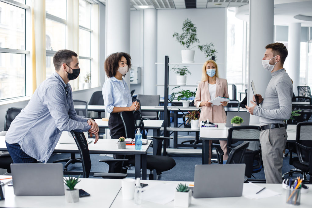 Modern team meeting, group work and social distancing. Manager with tablet speaks with workers in protective masks in interior of modern office with gadgets during coronavirus epidemic, free space